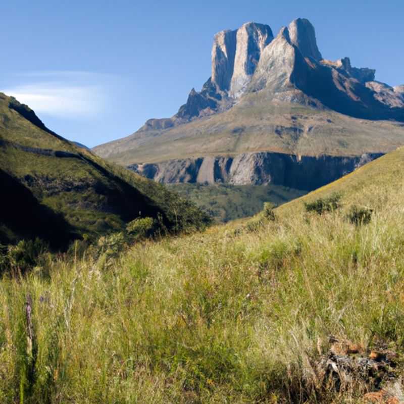 A solitary traveler stands dwarfed against the majestic Cathedral Peak, South Africa, its sheer rock faces reaching towards the heavens, during a three-week springtime adventure.